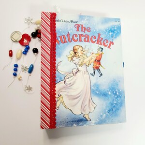 Peut inclure: Un exemplaire vintage du livre "The Nutcracker" de Little Golden Book, avec une illustration de couverture d'une femme en robe blanche tenant un soldat de plomb. La tranche du livre est décorée de papier à rayures rouges et blanches et d'une bordure en dentelle rouge. Des perles et des boutons décoratifs sont disposés à proximité.