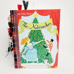 Peut inclure: Un journal fait à la main avec une couverture du livre d'or "The Nutcracker". L'illustration de la couverture représente une ballerine, un soldat et un sapin de Noël. La tranche est reliée avec un ruban à carreaux rouge et ornée de perles et de breloques.