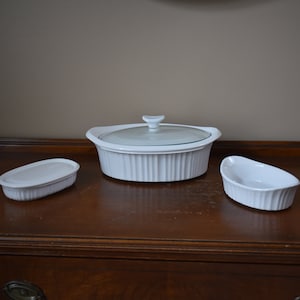 May include: A set of three white ceramic baking dishes with a glass lid. The dishes are oval shaped and have a ribbed design.