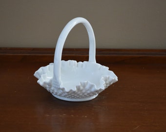 May include: White milk glass basket with a ruffled edge and a handle. The basket is empty and sitting on a wooden surface.