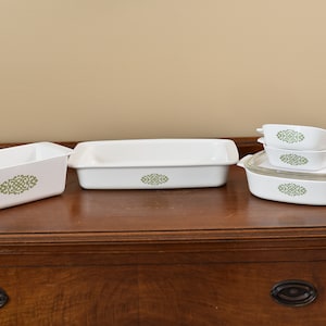 May include: A set of white baking dishes with a green floral design. The set includes a loaf pan, a rectangular baking dish, and three smaller dishes with lids.