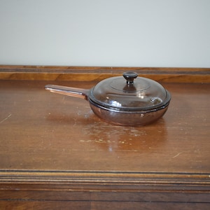 May include: A brown glass saucepan with a lid and a black handle. The saucepan is sitting on a wooden surface.