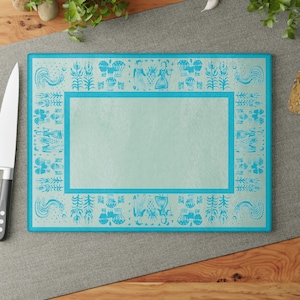 May include: A blue glass cutting board with a white design of stylized figures, plants, and mushrooms around the border. The center of the board is a light blue.