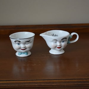 May include: Two white ceramic cups with winking faces. The larger cup has a handle and says "Yum" on the front. The smaller cup has a blue bow tie and says "Bailey's" on the front.