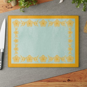 May include: A yellow and blue glass cutting board with a floral border. The board is rectangular and has a smooth surface.