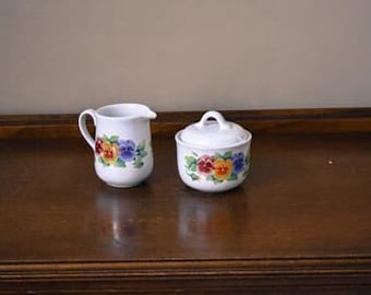 May include: White ceramic creamer and sugar bowl set with a floral design of yellow, red, blue, and purple pansies.