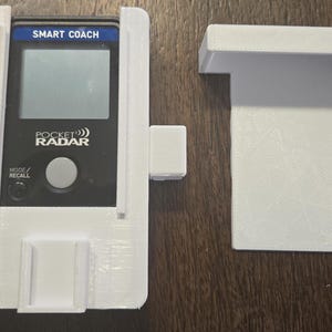 May include: A white 3D-printed holder for a Pocket Radar device. The holder has a rectangular shape with a slot for the device and a separate T-shaped piece. The device displays "SMART COACH" and "POCKET RADAR".