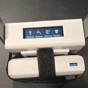 May include: A white electronic device with two gray antennas and a small screen displaying icons and text. The text on the screen reads "Ethernet Repeater Tethering Cellular". A black strap secures a white power bank to the device.