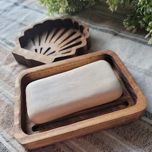 Op de afbeelding: Een houten zeepbakje met een schelpdesign. Het bakje is gemaakt van natuurlijk hout en heeft een rechthoekige vorm. Een stuk zeep ligt in het bakje.
