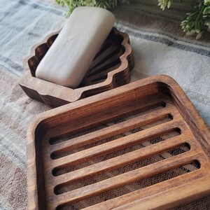 May include: Two wooden soap dishes. One is shaped like a seashell and the other is a rectangular dish with a slatted design. A bar of white soap is in the seashell dish.