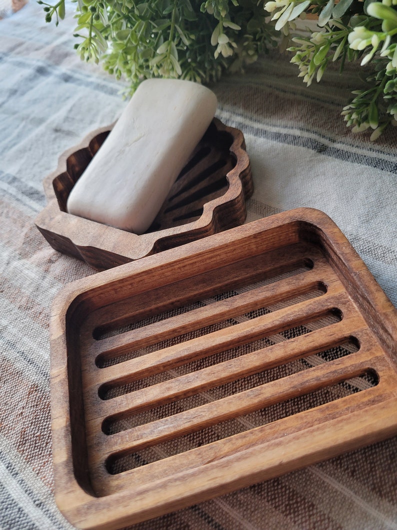 May include: Two wooden soap dishes. One is shaped like a seashell and the other is a rectangular dish with a slatted design. A bar of white soap is in the seashell dish.