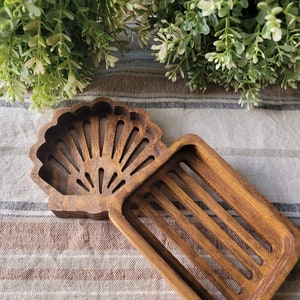 May include: A wooden soap dish with a seashell design. The dish has a rectangular shape with a raised edge and a cutout in the shape of a seashell. The seashell cutout has a fan-like design with multiple lines radiating from the center.
