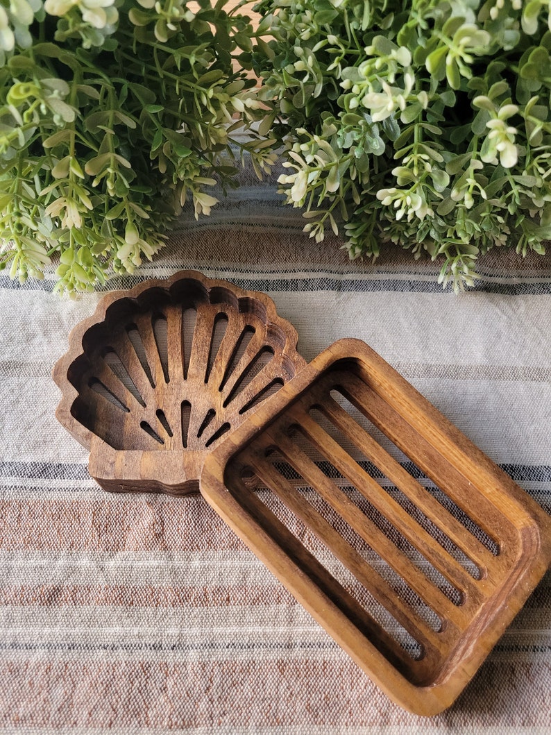 May include: A wooden soap dish with a seashell design. The dish has a rectangular shape with a raised edge and a cutout in the shape of a seashell. The seashell cutout has a fan-like design with multiple lines radiating from the center.