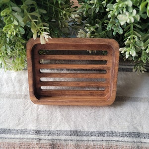 May include: A wooden soap dish with a slatted design. The dish is made of dark brown wood and has a rectangular shape.