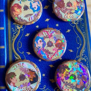 May include: Five round, holographic buttons featuring characters from the animated series Arcane. Each button has a different character pair, including Jinx & Vi, Powder & Ekko,  Jayce & Viktor, Silco & Vander, and  Jinx & Vi. The buttons are all gold with a holographic finish.
