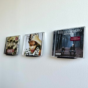 May include: Three CD cases are mounted on a white wall. The case on the left features a band with the text "Sunrise" on the cover. The middle case features a woman with a red lip and a black and white patterned shirt. The case on the right features the text "The Offspring Days Go By" on the cover.