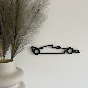 Peut inclure: Silhouette noire en métal d'une voiture de course montée sur un mur blanc. La voiture est orientée vers la droite et a un design simple.