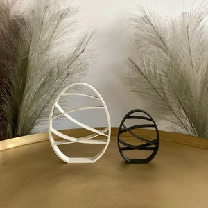 Op de afbeelding: Twee decoratieve paaseieren, een wit en een zwart, met een geometrisch ontwerp van gebogen lijnen. De eieren liggen op een gouden oppervlak.