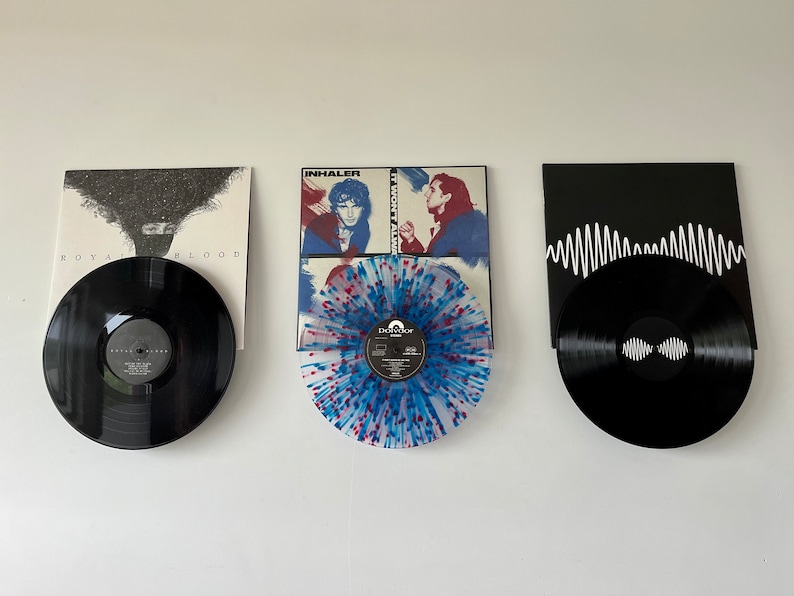 Peut inclure: Trois disques vinyles affich&eacute;s sur un mur blanc. Le disque de gauche est noir avec une &eacute;tiquette blanche et le texte "Royal Blood". Le disque du milieu est un vinyle splatter bleu, rouge et blanc avec une &eacute;tiquette noire et le texte "Dry Cleaning". Le disque de droite est noir avec une &eacute;tiquette blanche et le texte "AM" en ligne ondul&eacute;e.