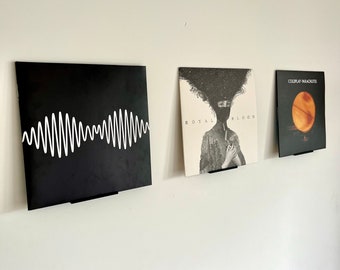 May include: Three black vinyl records are displayed on a white wall using black metal record holders. The record covers feature a white waveform pattern, a black and white image of a person with a black mask, and a red and orange image of a planet.