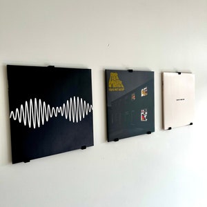 May include: Three vinyl records displayed on a white wall using black record holders. The record on the left has a black background with a white waveform pattern. The middle record has a black background with a yellow and white graphic. The record on the right has a white background.
