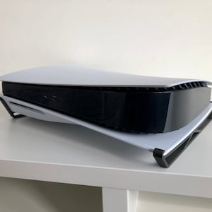 Op de afbeelding: Een witte PlayStation 5 console met een zwart voorpaneel staat op een wit rek. De console is lichtjes naar rechts gekanteld.