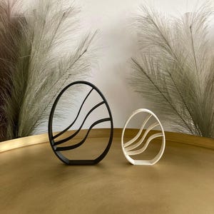 Può includere: Due tagliabiscotti decorativi a forma di uovo, uno nero e uno bianco, con un design ondulato. I tagliabiscotti sono su una superficie dorata.