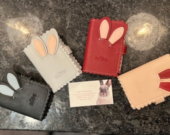 Puede incluir: Cuatro pequeñas carteras de imitación de cuero de colores con detalles de orejas de conejo. Cada cartera tiene un borde festoneado y el nombre de la marca "AnOther" en relieve en la parte delantera. Las carteras están dispuestas sobre una superficie gris con una tarjeta de visita que presenta un conejo en el centro.