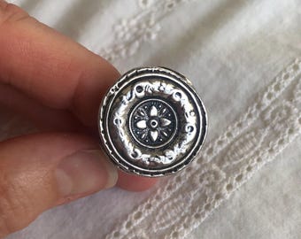 May include: A round silver-toned metal ring featuring a central floral motif surrounded by a textured, embossed border with swirling patterns. The surface of the metal has a darkened, oxidized finish in the recessed areas to highlight the details.