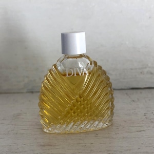 Op de afbeelding: Een kleine, heldere glazen parfumfles met een witte dop. De fles is gevuld met een gouden vloeistof en heeft het woord "DIVA" op de voorkant gedrukt. De fles heeft een gestructureerd, geribbeld ontwerp.
