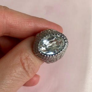 May include: A silver-toned ring featuring a large, oval-shaped clear faceted stone set in the centre. The band of the ring has a textured, woven-like metal pattern that surrounds the central stone, creating a detailed and reflective appearance.