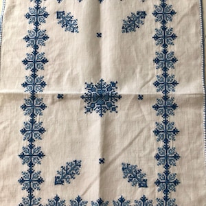 Op de afbeelding: Wit linnen tafelkleed met een gedetailleerd blauw geborduurd sneeuwvlokontwerp. De rand heeft een herhalend patroon van sneeuwvlokken, met extra sneeuwvlokclusters in het midden. Het borduurwerk is in een donkerdere blauwtint.