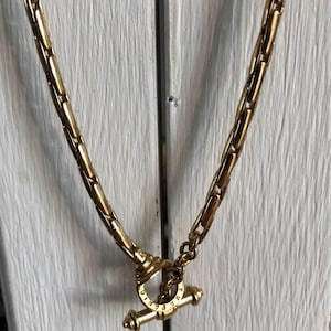 Pode incluir: Um colar de tom dourado com uma corrente espessa e texturizada composta por elos alongados e arredondados. O colar prende-se com um fecho de alavanca, consistindo num anel circular e uma barra em T, ambos em metal dourado polido.