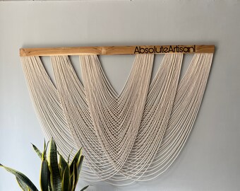 May include: A white macrame wall hanging with a wooden dowel at the top. The dowel has the text "AbsoluteArtisan" burned into it.