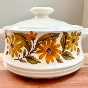 May include: White ceramic casserole dish with a floral design featuring yellow, orange, and green flowers. The dish has two handles and a lid.