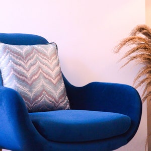 Pode incluir: Uma poltrona de veludo azul com uma almofada estampada. A poltrona está em uma sala com uma parede branca e um vaso de capim-dos-pampas seco.