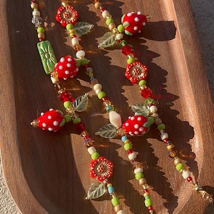 Peut inclure: Bijoux à thème fraise en perles. Les bijoux présentent des perles de fraises rouges à pois blancs, des perles de fleurs rouges, des perles de feuilles vertes et diverses perles colorées. Les bijoux sont présentés sur un plateau en bois.