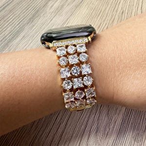 May include: A gold-tone smartwatch band featuring multiple rows of alternating square and round clear crystals. The band connects to a black smartwatch face, with a single row of small pavé crystals lining the attachment point to the watch case.