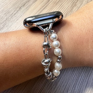 May include: A silver chain bracelet with a silver clasp and white pearl beads. The bracelet is attached to a black Apple Watch.