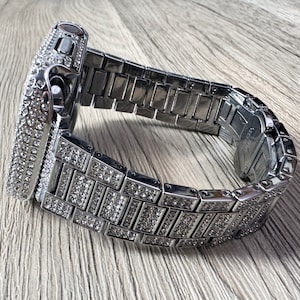Puede incluir: Una caja de reloj inteligente plateada y una pulsera de eslabones a juego, ambas completamente incrustadas con brillantes transparentes. El reloj presenta un marco rectangular con un bot&oacute;n lateral y un cierre met&aacute;lico seguro en la pulsera.
