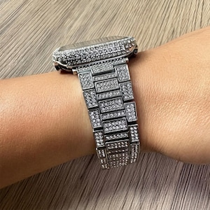 Puede incluir: Una caja de reloj inteligente plateada y una pulsera de eslabones a juego, ambas completamente incrustadas con numerosas piedras peque&ntilde;as, transparentes y brillantes. El reloj presenta una esfera rectangular y una correa de metal.