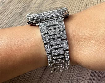 Puede incluir: Una caja de reloj inteligente plateada y una pulsera de eslabones a juego, ambas completamente incrustadas con numerosas piedras pequeñas, transparentes y brillantes. El reloj presenta una esfera rectangular y una correa de metal.