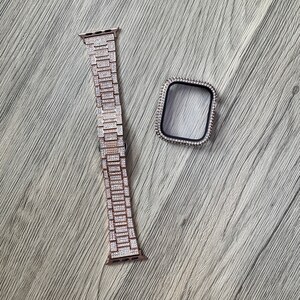 Puede incluir: Una correa de reloj inteligente en oro rosa y una funda protectora a juego, ambas completamente incrustadas con peque&ntilde;os cristales claros y brillantes. La correa presenta un dise&ntilde;o de eslabones met&aacute;licos y la funda rectangular tiene un borde interior negro.
