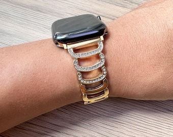 May include: Gold watch band with clear rhinestones. The band is made of metal and has a series of interlocking links. The band is designed to fit a smartwatch.