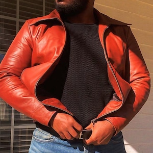 May include: A red leather jacket with a wide collar and a zip closure. The jacket is worn over a black sweater and blue jeans. The person is fastening the belt of the jacket.