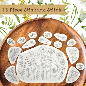 May include: A wooden tray displays 15 stick-and-stitch embroidery pieces. The pieces feature bee and floral designs. The text "15 Piece Stick and Stitch" is at the top. The background is white with floral and leaf patterns.