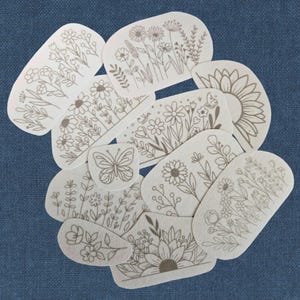 K&ouml;nnte beinhalten: Eine Ansammlung von cremefarbenen, ovalen Aufn&auml;hern mit braunen Strichzeichnungen von Blumen und Pflanzen, darunter Sonnenblumen und ein Schmetterling. Die Aufn&auml;her sind auf einem blauen Stoffhintergrund angeordnet. Ma&szlig;e nicht zutreffend.
