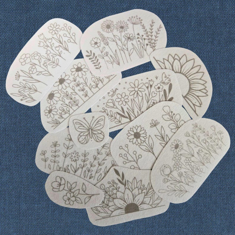 K&ouml;nnte beinhalten: Eine Ansammlung von cremefarbenen, ovalen Aufn&auml;hern mit braunen Strichzeichnungen von Blumen und Pflanzen, darunter Sonnenblumen und ein Schmetterling. Die Aufn&auml;her sind auf einem blauen Stoffhintergrund angeordnet. Ma&szlig;e nicht zutreffend.