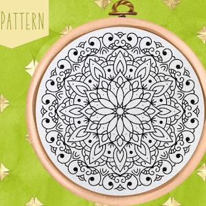 Op de afbeelding: Een zwart-wit mandala-patroon in een ronde borduurring. De ring is gemaakt van hout en heeft een lichtbruine kleur. De achtergrond is een groen en goud patroon.