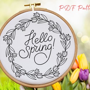Puede incluir: Un patrón de bordado en blanco y negro de una corona floral con el texto "Hello Spring!" en su interior. El patrón se muestra en un aro de bordado de madera.